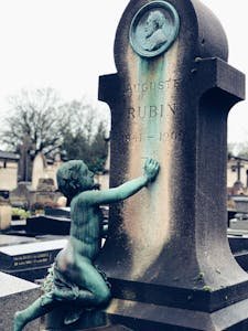 A detailed view of a bronze headstone cherub sculpture adorning Auguste Rubin's headstone in a Parisian cemetery.