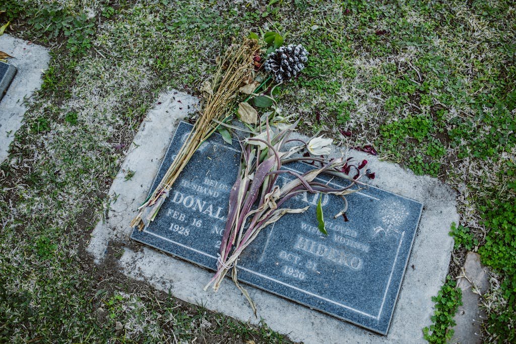 A gravestone with dried flowers lying on it in a cemetery, featuring grass and engraved details. What to put on grave before headstone.