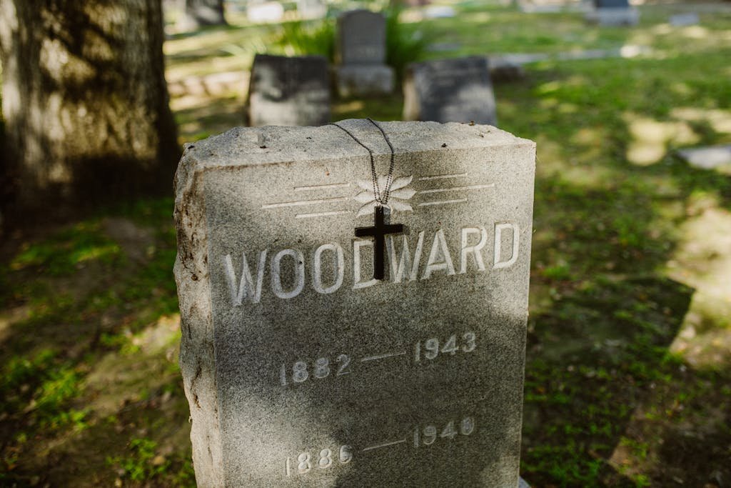 A peaceful cemetery scene with a gravestone marked 'Woodward' and a cross necklace hanging in sunlight. What to have written on a headstone.