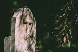 Close-up of a weathered tombstone with a cross in an overgrown cemetery, casting shadows.