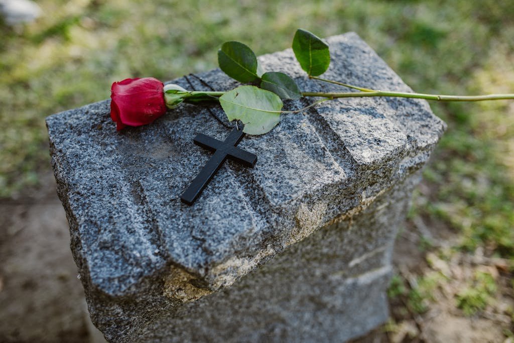 A red rose and black cross placed on a gray granite gravestone in a serene outdoor setting.