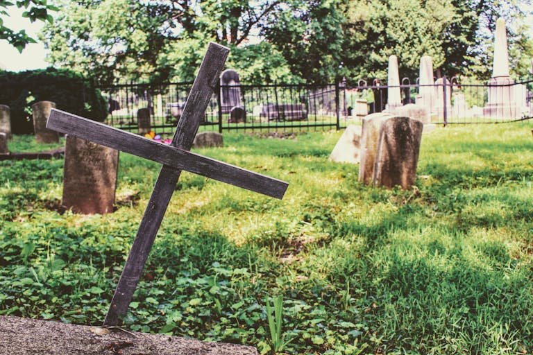 A serene cemetery with a wooden cross and tombstones on a sunny day, surrounded by nature. Can you be buried without a casket.