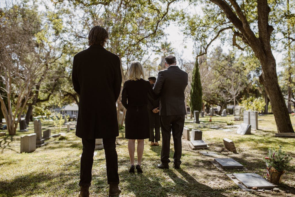 A solemn gathering of adults in formal attire at a serene cemetery setting, beneath trees.