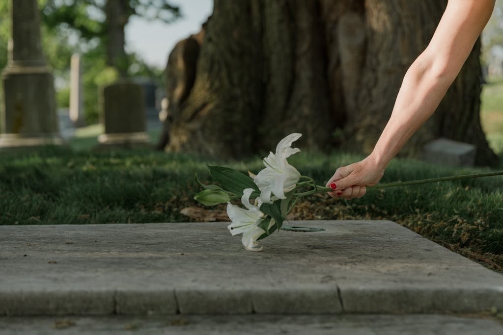 A solemn moment of placing white lilies on a cemetery gravestone, symbolizing loss and remembrance. What to Bring When Visiting a Grave.
