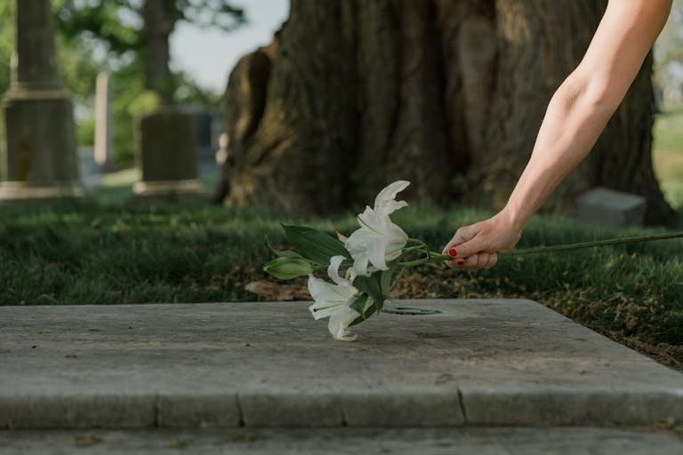 A solemn moment of placing white lilies on a cemetery gravestone, symbolizing loss and remembrance. What to Bring When Visiting a Grave.