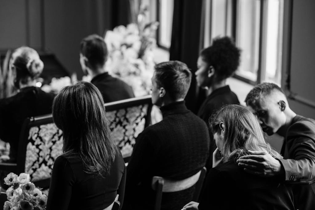 A somber grayscale image of mourners sitting indoors during a funeral, conveying loss and sympathy.