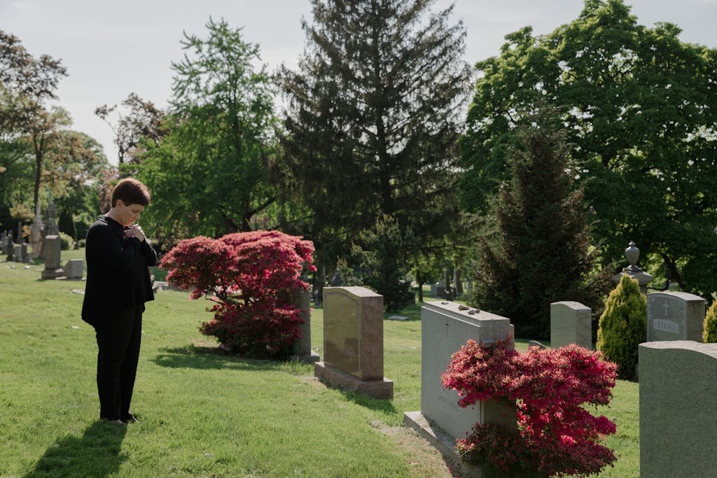 A woman stands thoughtfully in a picturesque cemetery, surrounded by green trees and vibrant flowers. Visiting a grave.