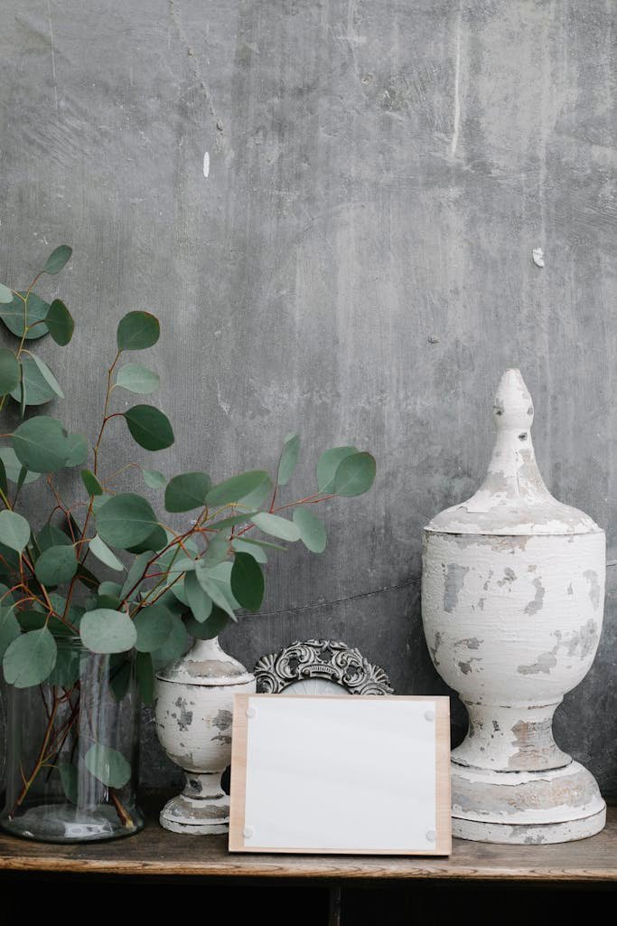 Board with blank paper sheet placed near shabby retro decorations and plant twigs against gray wall