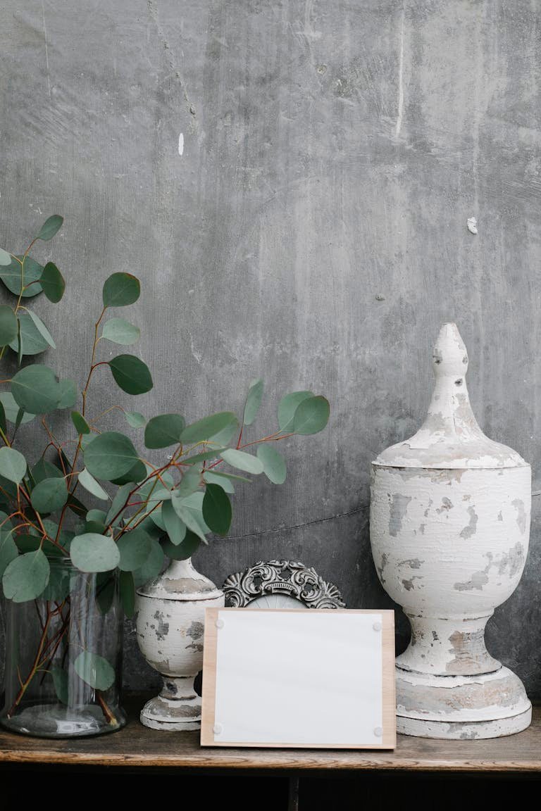 Board with blank paper sheet placed near shabby retro decorations and plant twigs against gray wall